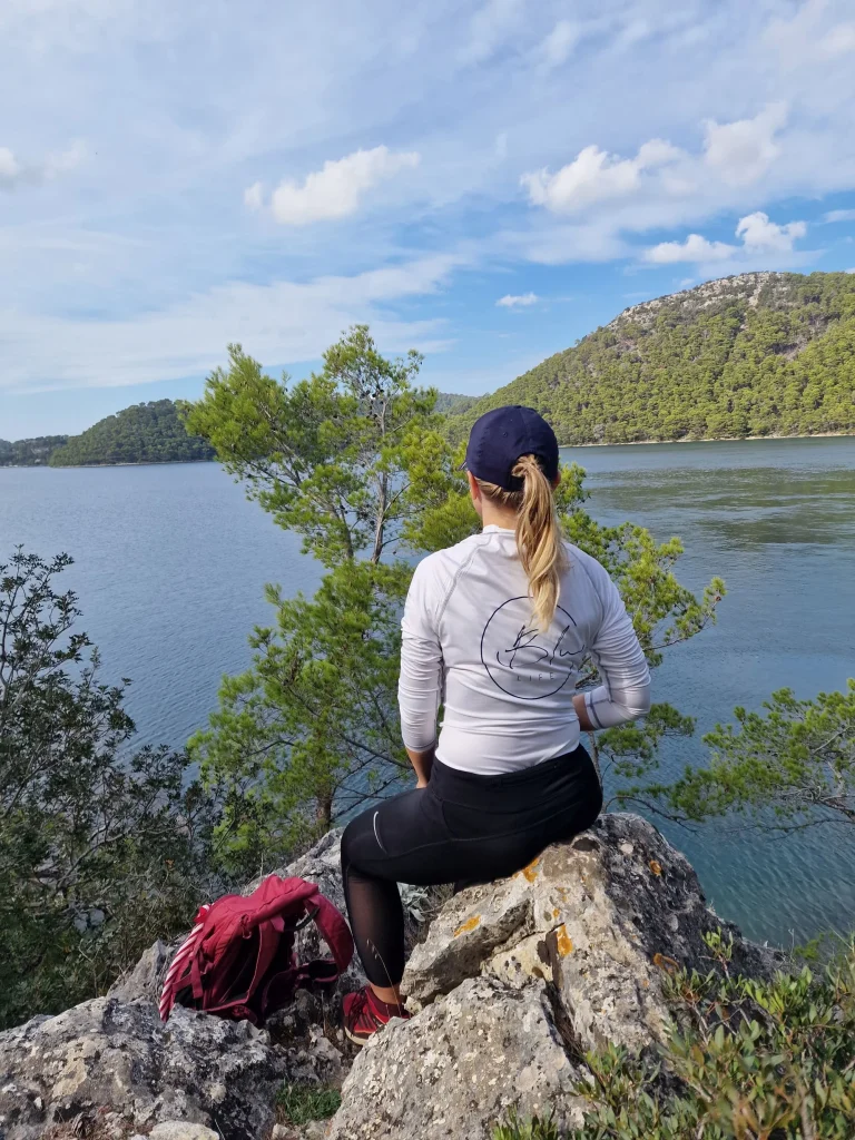 A person in outdoor gear, wearing a cap, sits on a rock overlooking a scenic lake surrounded by lush green hills. The sky is partly cloudy. A red backpack lies on the rocks beside them.